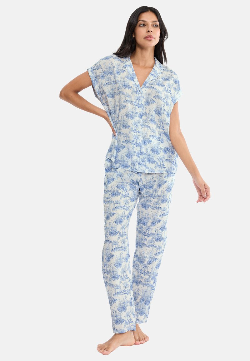 Kobieta stoi boso ubrana w dopasowany, luźny zestaw piżam z krótkim rękawem i spodniami w niebiesko-biały wzór kwiatowy.