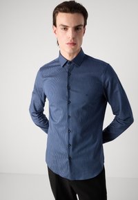 Cămașă bleumarin cu nasturi, cu dungi verticale fine, mâneci lungi și guler clasic. Prezintă nasturi negri și un logo subtil pe piept.