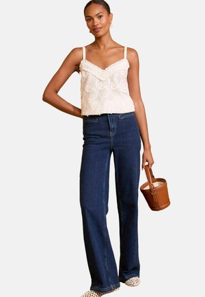 Woman wearing white lace sleeveless top, dark blue high-waist jeans, white woven shoes, holding a brown bucket bag with a handle.
