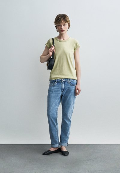 Νεαρός/νεαρή στέκεται μπροστά σε ένα απλό τοίχο, φορά ένα ανοιχτό πράσινο t-shirt, μπλε jeans με γυρισμένα μπατζάκια, μαύρες φλατ μπότες και κρατά μια μικρή μαύρη τσάντα ώμου.