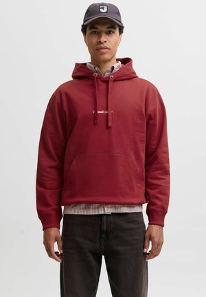 Man wearing a red hoodie over a checkered shirt, black jeans, and a black baseball cap standing against a plain light background.