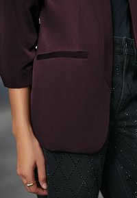 Primo piano di una persona che indossa un blazer viola scuro con tasca e pantaloni neri con motivi a forma di diamante, mano rilassata al fianco.