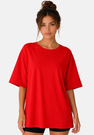 Jeune femme aux cheveux bruns en chignon, portant un T-shirt rouge uni et ample et un short noir, debout sur un fond blanc.