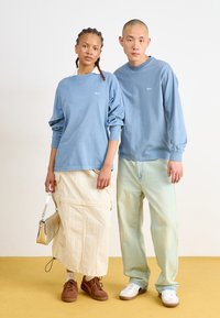 Light blue long-sleeve shirts with a small logo, beige skirt, light green jeans, brown and white sneakers, and silver handbag on a yellow floor.