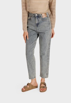Jeans in denim chiaro con vestibilità rilassata, caratterizzati da un alto punto vita, leggero sbiadimento e dettagli di cucitura laterale; abbinati a sandali marroni.