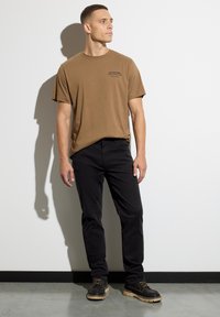 Mężczyzna w brązowej koszulce z nadrukiem, czarnych spodniach i czarnych butach stoi pod białą ścianą, patrząc w bok.