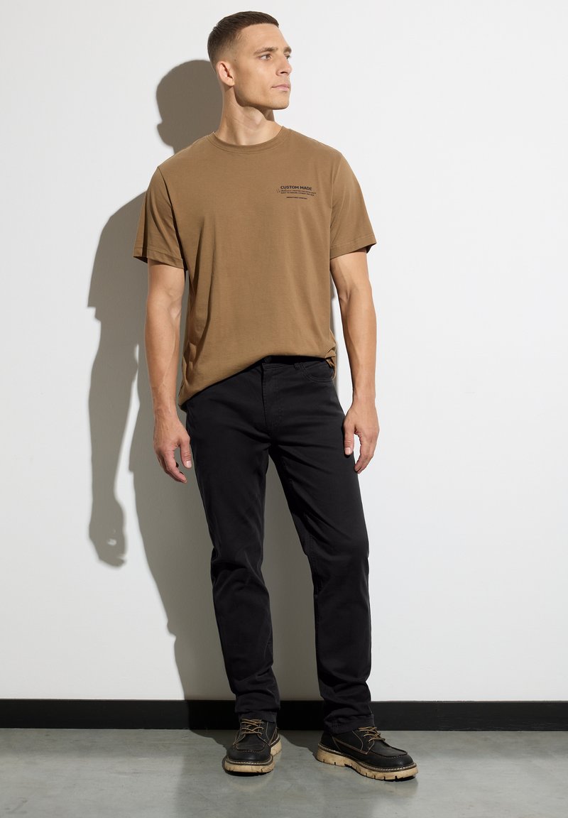 Mężczyzna w brązowej koszulce z nadrukiem, czarnych spodniach i czarnych butach stoi pod białą ścianą, patrząc w bok.