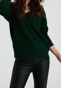 Dunkelgrüner Strickpullover mit V-Ausschnitt, gerippten Bündchen und Saum. Kombiniert mit schwarzen, eng anliegenden Hosen wirkt die Textur weich und gemütlich.