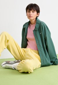 Grøn letvægtsjakke, lyserød tætsiddende top og gule sweatpants. Hvide sneakers med reflekterende accenter. Sidder på grøn overflade.