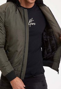 Giacca bomber verde oliva con interno nero, dotata di chiusura a zip e polsini a costine, indossata sopra una maglietta nera con testo bianco.