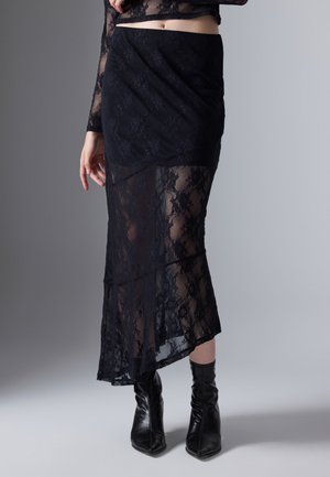 Kvinde iført en sort blondernederdel med gennemsigtige paneler og sorte højhælede ankelstøvler, stående på en ensfarvet lys baggrund.