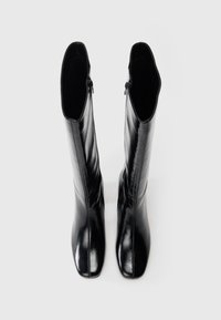 Spīguļojošu melnu ādas zābaku pāris līdz ceļiem ar kvadrātveida purngalu un sānu rāvējslēdzējiem, skatīts no augšas uz balta fona.