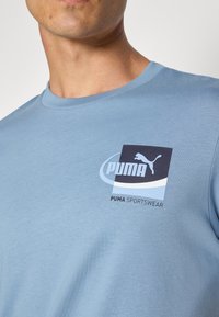 Tricou din bumbac, de culoare albastru deschis, cu mânecă scurtă, cu un decolteu rotund și un logo grafic mare care prezintă un puma sărind în gri închis.