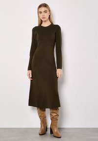 Strickkleid mit langen Ärmeln in Olivgrün, runder Ausschnitt, tailliertem Oberteil und fließendem Rock, kombiniert mit beigen, knielangen Stiefeln mit Absatz.