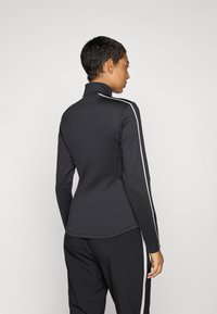 Fekete sportos dzseki magas gallérral, fehér oldalsávokkal, testhez simuló fazonnal, sima textúrával és finoman ívelt szegéllyel.