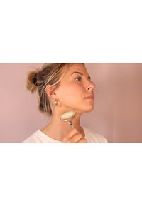 Jade ansiktsroller hålls av en person med ljus hud, med ett metallhandtag och ett rundat stenhuvud, mot en mjuk rosa bakgrund.