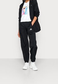 Zwarte windbreakerjack over een wit T-shirt met een kleurrafische afbeelding, gecombineerd met zwarte joggerbroeken en witte sneakers.