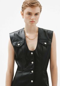 Vestido de cuero negro con un profundo escote en V, diseño sin mangas, botones a presión en la parte delantera y dos bolsillos en el pecho. Acento de cadena dorada.