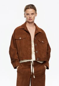 Giacca cropped in suede marrone con collo, bottoni a pressione argentati e due tasche sul petto. Indossata sopra un top crema, che mette in risalto una texture morbida.