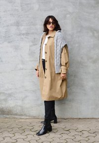 Béžový oversize trenčkot s veľkými gombíkmi, viacvrstvový sivý pletený šál, čierne priliehavé džínsy a členkové čižmy. Hladká štruktúra a neutrálné farby.