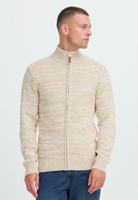 Mann iført en beige strikkegenser med høy krage og glidelås, ser mot høyre, kombinert med blå jeans mot en ensfarget bakgrunn.