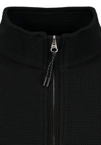 Čierny odev so vzorom vafľovej pleteniny so strieborným zipsom a čiernymi ťahacími šnúrkami, so stojacím, štruktúrovaným golierom.