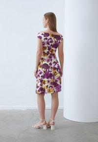 Blumenmusterkleid mit lila, gelben und weißen Mustern, im Off-Shoulder-Design und in knieumspielender A-Linie. Flache Sandalen runden den Look ab.