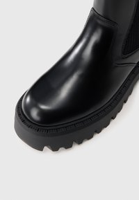 Bota de tornozelo preta em borracha com superfície brilhante, painéis laterais elásticos e uma sola espessa e padronizada com cravos profundos para tração.