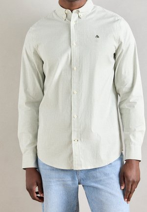 Homme portant une chemise boutonnée à manches longues à fines rayures vert clair et blanc avec un jean bleu clair sur un fond uni.