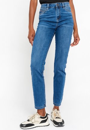 Femme portant un jean slim bleu et des baskets noir et blanc, debout devant un fond blanc.
