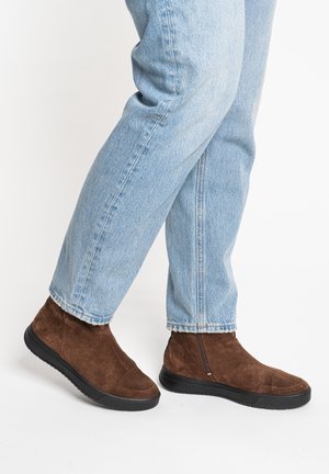 Beine, die hellblaue Jeans und braune Wildleder-Stiefeletten mit schwarzen Sohlen tragen, stehen auf weißem Hintergrund.