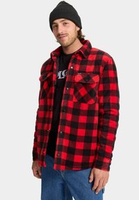 Camisa de franela a cuadros roja y negra, con botones a presión, dos bolsillos en el pecho y una camiseta negra debajo, combinada con vaqueros azules.