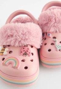 Zuecos rosas con un puño de piel de peluche rosa, que cuentan con acentos de goma decorativos en forma de abeja, mariquita, arcoíris y dinosaurio. Superficie texturizada.