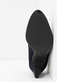 Chaussure noire à bout pointu et talon haut, posée sur son talon, montrant une semelle propre et une petite pointe de talon sur une surface blanche.