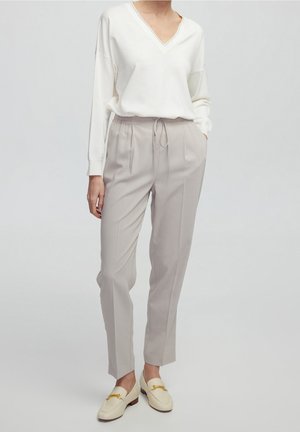 Woman wearing white V-neck sweater, light beige drawstring trousers, and cream-colored loafers with gold buckles, standing against plain background.