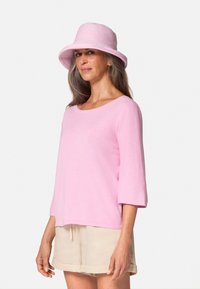 Lichtroze pullover bovenstuk met driekwart mouwen, gecombineerd met beige shorts; zachte textuur. Accessoire: lichtroze hoed met brede rand.