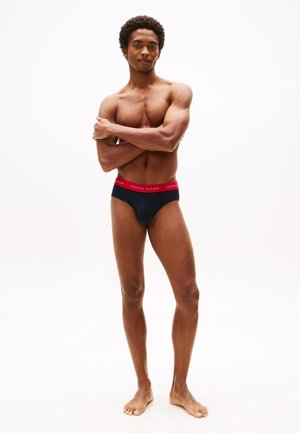 Marineblaue Slips mit einem roten elastischen Bund, der das Logo "TOMMY HILFIGER" trägt. Das Design ist enganliegend und aus einem glatten Stoff gefertigt.