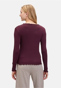 Haut à manches longues en tissu côtelé violet profond avec des bords festonnés au niveau du col et des poignets, associé à un pantalon gris clair texturé.