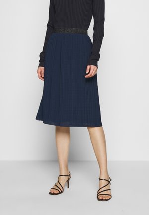 Femme portant une jupe midi plissée bleu marine avec une ceinture noire pailletée, associée à un haut noir à manches longues et des sandales noires à talons hauts à brides.