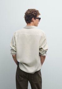 Lichtbeige shirt met opgerolde mouwen, klassieke kraag, relaxte pasvorm en gestikte details op de rug, gecombineerd met olijfgroene broeken.