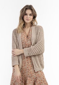 Beige gebreide cardigan met open weving, geribde manchetten en zoom, gedragen over een bloemenjurk met een ruches rok en ronde halslijn.