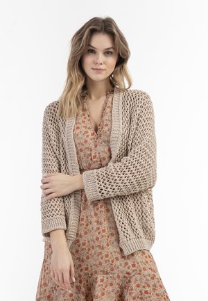 Beige gebreide cardigan met open weving, geribde manchetten en zoom, gedragen over een bloemenjurk met een ruches rok en ronde halslijn.