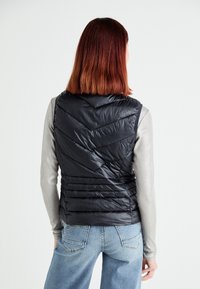 Gilet rembourré noir avec un motif en chevrons, texture brillante et col haut, porté sur un haut à manches longues gris et associé à un jean bleu.