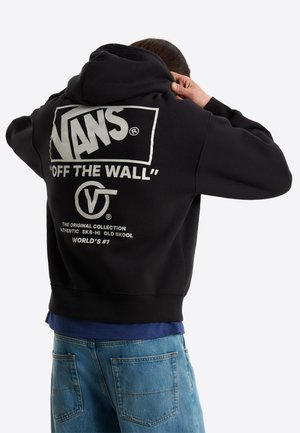 Personne portant un sweat à capuche noir Vans avec le texte « Off The Wall » et un logo dans le dos, une chemise bleue en dessous et un jean bleu en denim.