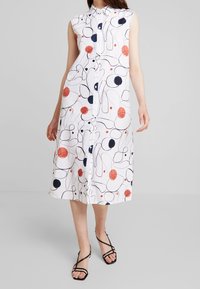 Abito bianco senza maniche con motivo astratto blu navy e rosso, lunghezza al ginocchio, design con bottoni davanti e tessuto leggero. Sandali neri con cinturini.