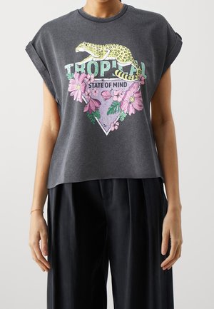 Mujer con una camiseta sin mangas gris oscuro con las mangas remangadas, con motivos florales y de leopardo, y texto que dice "Tropical State of Mind."