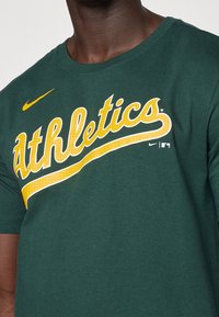 Tmavě zelené bavlněné tričko s velkým žlutým nápisem "Athletics" ve zkoseném, tučném písmu s jemnou texturou a logem Nike na hrudi.