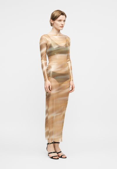 Γυναίκα όρθια, φοράει ένα διάφανο, μακρυμάνικο, μπεζ φόρεμα μήκους μέχρι το πάτωμα πάνω από ταιριαστό σουτιέν και εσώρουχα, συνδυασμένο με μαύρα πέδιλα με λουράκια.