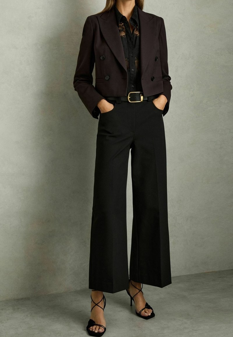 Femme portant un blazer noir court croisé, un chemisier noir à fleurs, un pantalon noir taille haute à jambes larges et des talons noirs à brides.