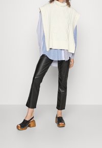 Krémový bezrukávový svetr s pleteným vzorem přes modro-bíle pruhovanou košili, s černými kalhotami z umělé kůže a černými kloboukovými botami na podpatku se dřevěnými patami.